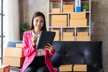 Young woman, an Asian online business owner, smiles, prepares parcel boxes and checks online orders...