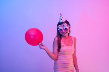 A young female birthday celebrant wearing a party hat and novelty glasses and holding a red...