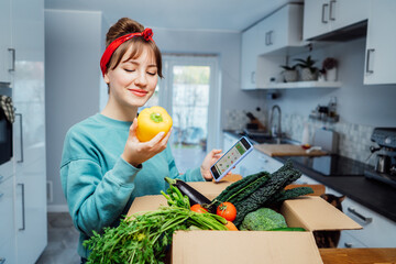 Online home food delivery. Woman checking her online order list on her phone. Cardboard box with fresh vegetables and fruits standing on the kitchen table. Local farmer food. Start of a healthy life