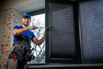 Builder in uniform repairs a plastic window with a screwdriver indoors. 