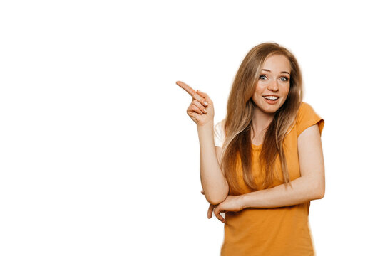 Cute cheerful redhead young woman wears orange casual clothes pointing aside at empty space by index finger, smiles broadly, discount time is on, over transparent background. Happy people concept.