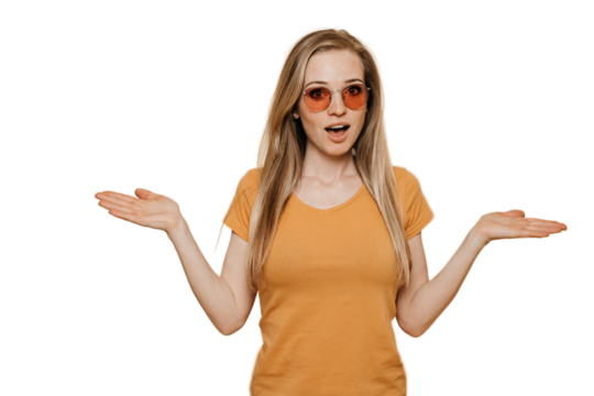 Shocked young red haired woman in sunglasses, dressed in orange t-shirt, spreads her hands in surprise, making choice, posing over transparent background. People sincere emotions.