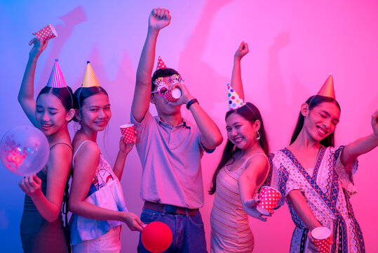 Five revelers holding cups and wearing party hats dancing and drinking alcohol. People and plain background lit with pink and blue lights.