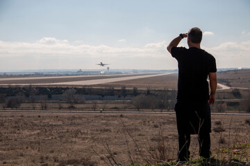 Slightly bald adult man dressed in black watching and pointing as planes take off from the airport runway
