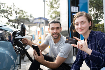 Young couple holding black credit card an electric car charging station.