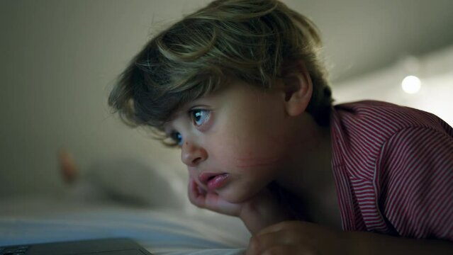 Child watching entertainment media on laptop screen laying in bed at night. Hypnotized kid staring at blue glowing screen