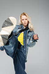 young blonde model in stylish denim outfit and trendy sneakers kick posing isolated on grey.