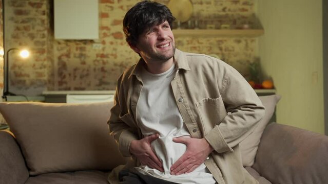 Upset unhealthy young man, sitting on the sofa in the living room, holding his stomach, suffering from cramp, stomach pain, poisoning, needs treatment. 