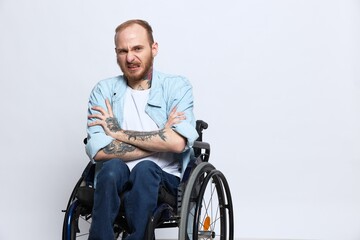 A man in a wheelchair looks at the camera anger and aggression, with tattoos on his arms sits on a gray studio background, health concept man with disabilities