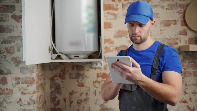 The technician servicing the water heater. A man checks the boiler room equipment using a tablet. 