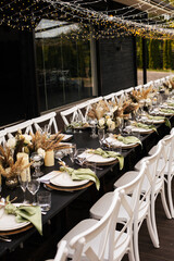 Wedding decorations. Served wedding table with white chairs golden plates, green napkins, decorative fresh and dried flowers, candles and and light bulbs. Celebration details, wedding outdoor
