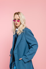 young blonde woman in blue blazer with tie and sunglasses posing with hands in pockets isolated on pink.