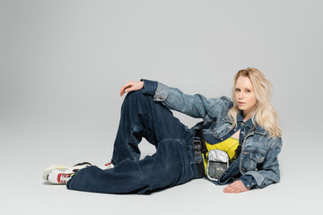 full length of young blonde woman in blue denim outfit and trendy sneakers posing on grey background.