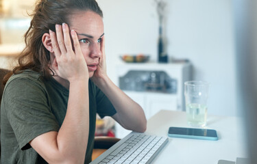 Surprised woman holding head with her hands looking at screen of computer while sitting at desk.
