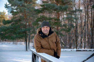 A man in the park in winter stands leaning on the railing against the backdrop of green pines