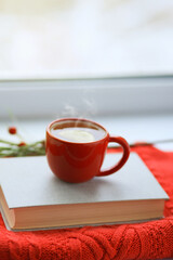 A cup of tea with a slice of lemon remains on a stack of knitted textile sweater on the window with a close-up book. The winter cozy season. Selective focus