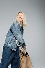 Fototapeta premium happy young woman in stylish denim outfit holding shopping bags and posing isolated on grey.