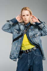 young blonde woman in stylish denim outfit looking at camera while posing with hands behind head...