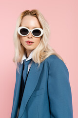 young blonde woman in blue blazer and sunglasses looking at camera isolated on pink.