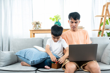 An Asian single father watches his son do his homework on the sofa in the living room of the house. While father works on a laptop computer. work from home online