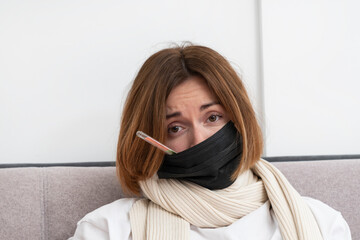 Young upset woman in medical mask sitting in the bed and measuring her fever. Isolation and pandemic concept	