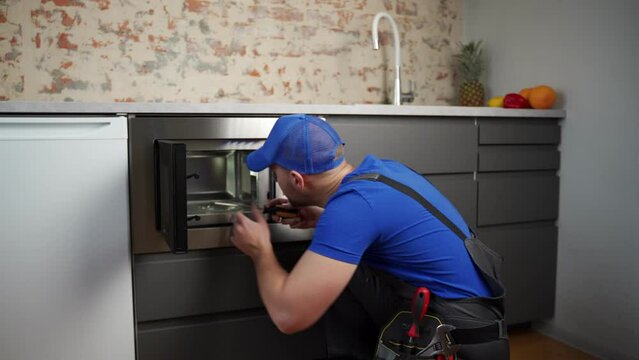 Young repairman in a blue uniform repairs and repairs a microwave oven with a screwdriver in a modern kitchen. 