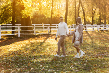 Fototapeta premium Young couple, man and pregnant wife are holding hands and walking in the autumn park on sunset. Happy family enjoying pregnancy on nature. The concept of Mother's Day and Women's Day. copy space.