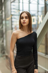 Stylish beautiful young girl model in fashionable black clothes with a one-shoulder top in an office building