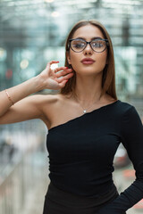 Fashion beautiful successful young business girl with glasses in a black sexy top stands and poses in a modern glass office building