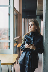 A smiling young woman is standing with a mobile phone in her hands and looking to the side while standing near a window reflecting sunlight. Modern workspace. Woman freelancer