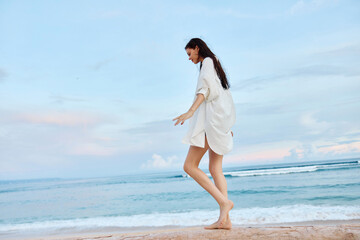 Happy tanned woman in white swimsuit shirt and denim shorts walks the beach on a log on the sand by the ocean with wet hair after swimming, sunset light in Bali