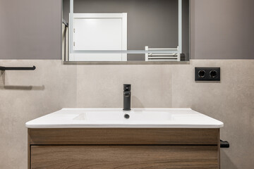 Square white ceramic sink on wooden table with drawers. On wall is mirror with white neon lighting. Mirror reflects white door and towel radiator. Walls are combination of marble tiles and gray paint.