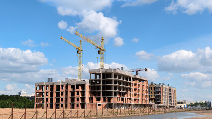 View of a construction site with multi-storey residential buildings under construction and several cranes against a blue sky with clouds.