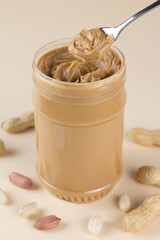 Fototapeta na wymiar Open jar with crunchy peanut butter, peanut and a spoon on a beige background. American food.