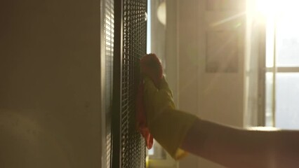 Closeup hands of unrecognizable cleaning service employee male in rubber gloves wiping radiator grill of refrigerator of collected dust and dirt using soft cleaning cloth in sunlight, slow motion.