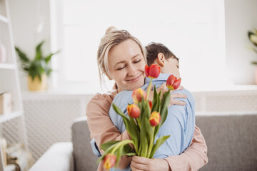 Cute boy sitting on the sofa with mom and giving a bouquet of tulips to her. - 572421389