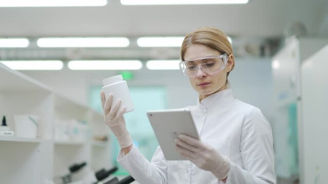 Focused mature woman scientist researcher with protective glasses looking at drug medicine product and compares the research in digital tablet at microbiology pharmaceutical biochemistry laboratory