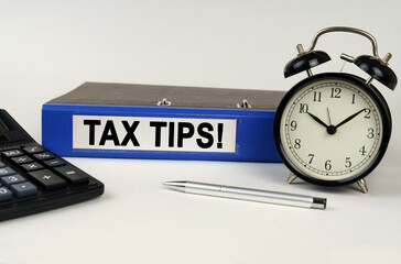 On a white surface, an alarm clock calculator and a folder with the inscription - Tax Tips