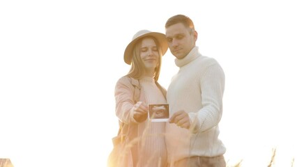 Happy young pregnant woman with husband are holding an ultrasound scan photo of the unborn child outdoors on nature in sunlights. Mothers day. A couple in love is waiting for the birth of a baby - Powered by Adobe