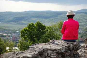 Fototapeta na wymiar A woman in a purple shirt sits on a rock with a river and mountains in the background.