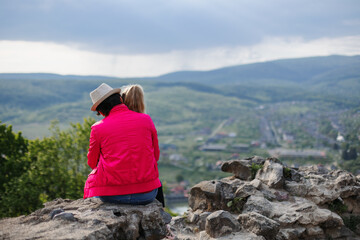 Naklejka premium A woman sitting on a rock in a purple jacket and hugging a child with a view of the river and mountains in the background.