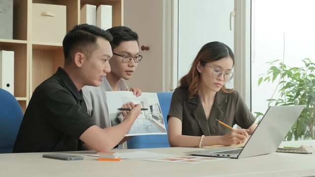 Team of young Asian coworkers discussing interior design plan via online video call on laptop while working together in office