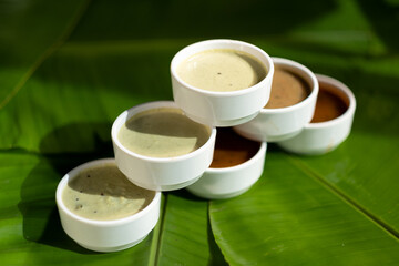 Showcased against a backdrop of banana leaves, several popular Indian sauces are served in small white dishes.