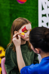 A professional make-up artist, artist paints with a brush on her face with face paints, drawing, children's makeup for a little girl model, a child. Photography, art, concept, lifestyle.