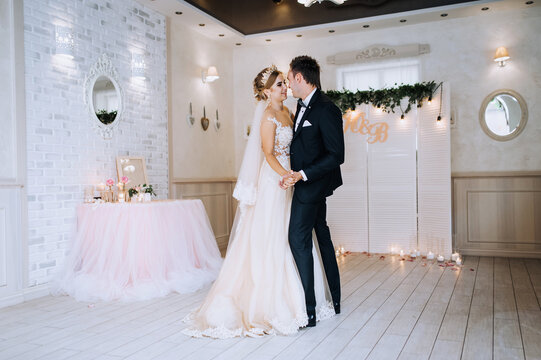 A young, stylish groom in a black suit and a beautiful blonde bride with a diadem in a dress are hugging, dancing in a restaurant, indoors. Wedding portrait, photo of happy newlyweds.