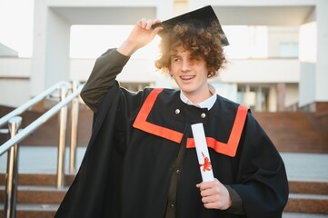 Low angle portrait of happy triumphant male graduate standing near university holding up diploma....