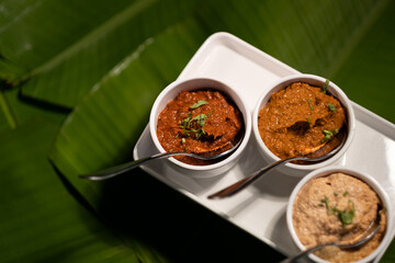 Three chutneys — ginger garlic chutney, tomato chutney, and peanut chutney — served fresh, against a backdrop of banana leaves.