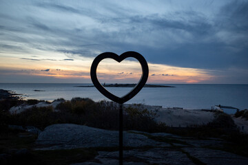 sculpture en forme de coeur évidé, au bord de l'ocean au coucher du soleil, on aperçoit une ile...