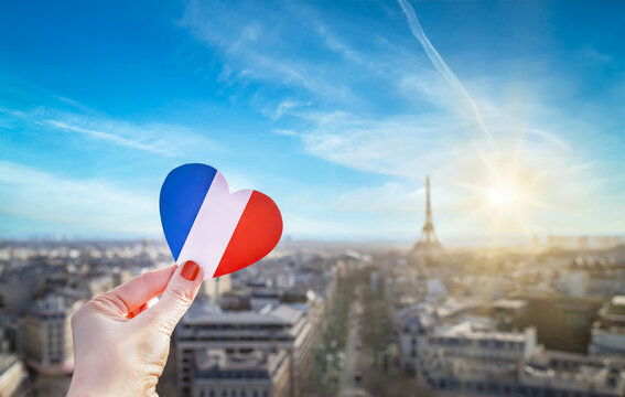 Girl's hand holding heart with french flag colors. Heart is on background of historic city center of Paris, Eiffel Tower, blue sky, clouds, sun. Love, Paris, travel concept.
