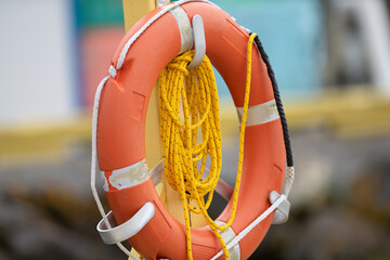 A vibrant orange polyethylene life ring hanging on a wooden post with long orange float line. There is reflective table on the preserver with white grab rope. The rescue equipment is a buoyant throw. 